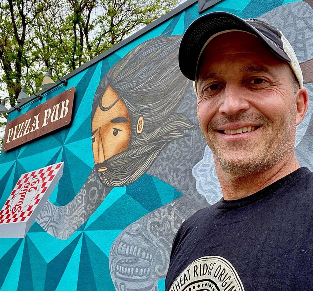 Mark Eskow, Owner of Right Coast Pizza in Wheat Ridge, stands in front of a mural
